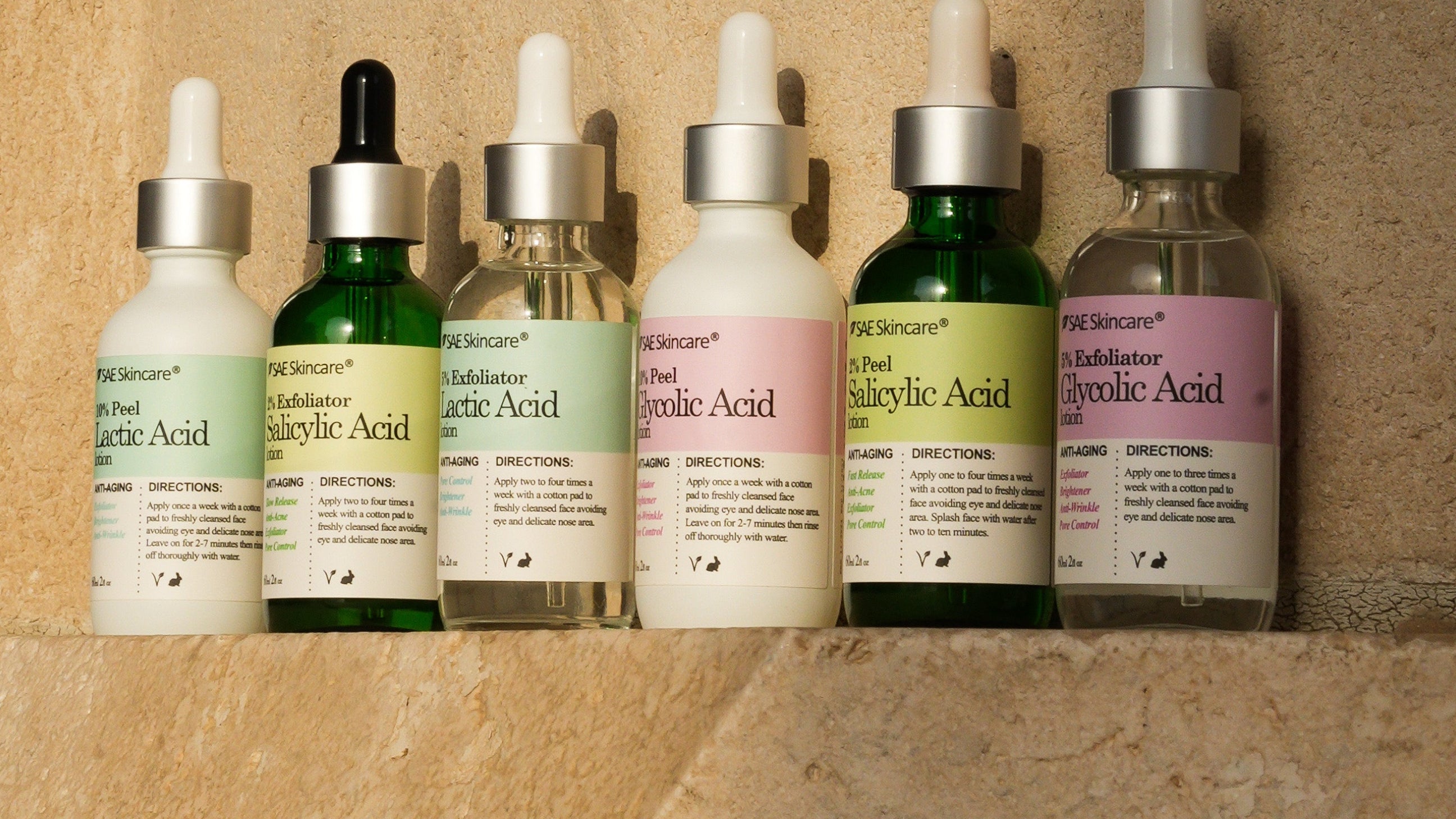Six skincare dropper bottles lined up on a beige stone surface, featuring white, green, and pastel pink labels with silver and black droppers.