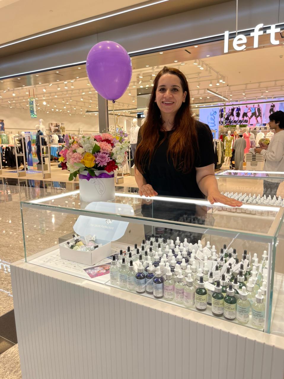 Sarah Saleem standing behind a counter with various bottles, a purple balloon, and flowers in a mall setting.